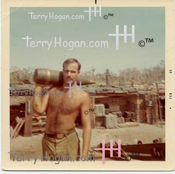 real-american-patriot-premium-rare-photograph-vietnam-gobart-humping-a-ninety-eight-pound-projo Crewmember Gobart walks toward a 155mm Howitzer, carrying a 98-pound fused explosive device on his shoulder. Behind him, a clear blue sky frames dense jungle foliage and a fortified ammunition cache protected by sandbags and wooden ammo boxes.