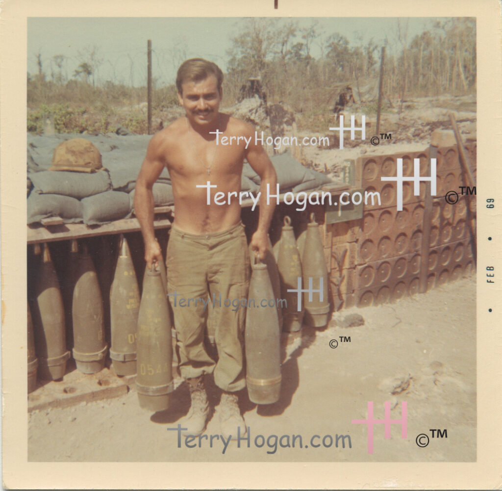 Pete stands holding an artillery projectile in each hand. Behind him are sandbags, upright projectiles, ammunition containers, a helmet, and a sign marked “P.”