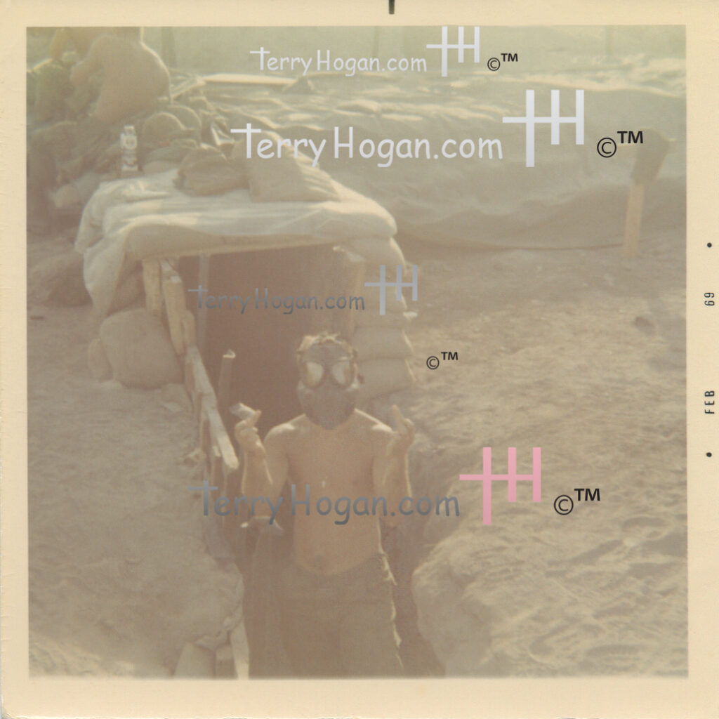 Pete wears a gas mask and raises both middle fingers in a narrow bunker entrance beneath stacked sandbags. The surrounding ground is bare, and the Polaroid’s right border reads “FEB 69.”
